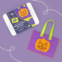A Halloween-themed felt bag kit shown with its printed packaging box on a lavender background. The kit includes a purple felt bag with a stitched orange jack-o'-lantern patch and bright green handles, alongside the box labeled 'Stitch Your Own Trick or Treat felt bag'.