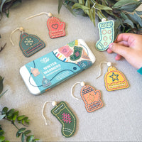 A tin of hand-embroidered wool felt Christmas ornaments is displayed on a neutral fabric surface with green foliage accents. A person's hand is holding one of the green stocking ornaments.