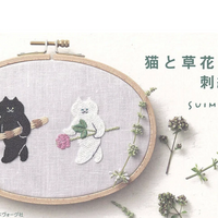 Embroidery hoop with black cat and white cat design on fabric, surrounded by small plants.