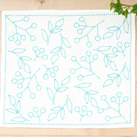 Embroidery pattern with blue thread on a white fabric square, placed on a wooden surface with green leaves.