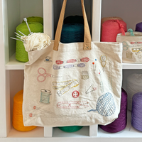Tote bag with craft illustrations hanging on a shelf with colorful yarn balls