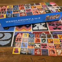 Puzzle box labeled 'Needlepoint A to Z' on a partially completed needlepoint canvas with alphabet and number patterns.