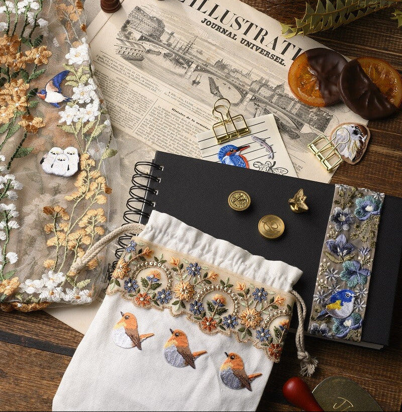Decorative items including a notebook, embroidered pouch, and candles on an illustrated newspaper.