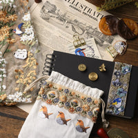 Decorative items including a notebook, embroidered pouch, and candles on an illustrated newspaper.