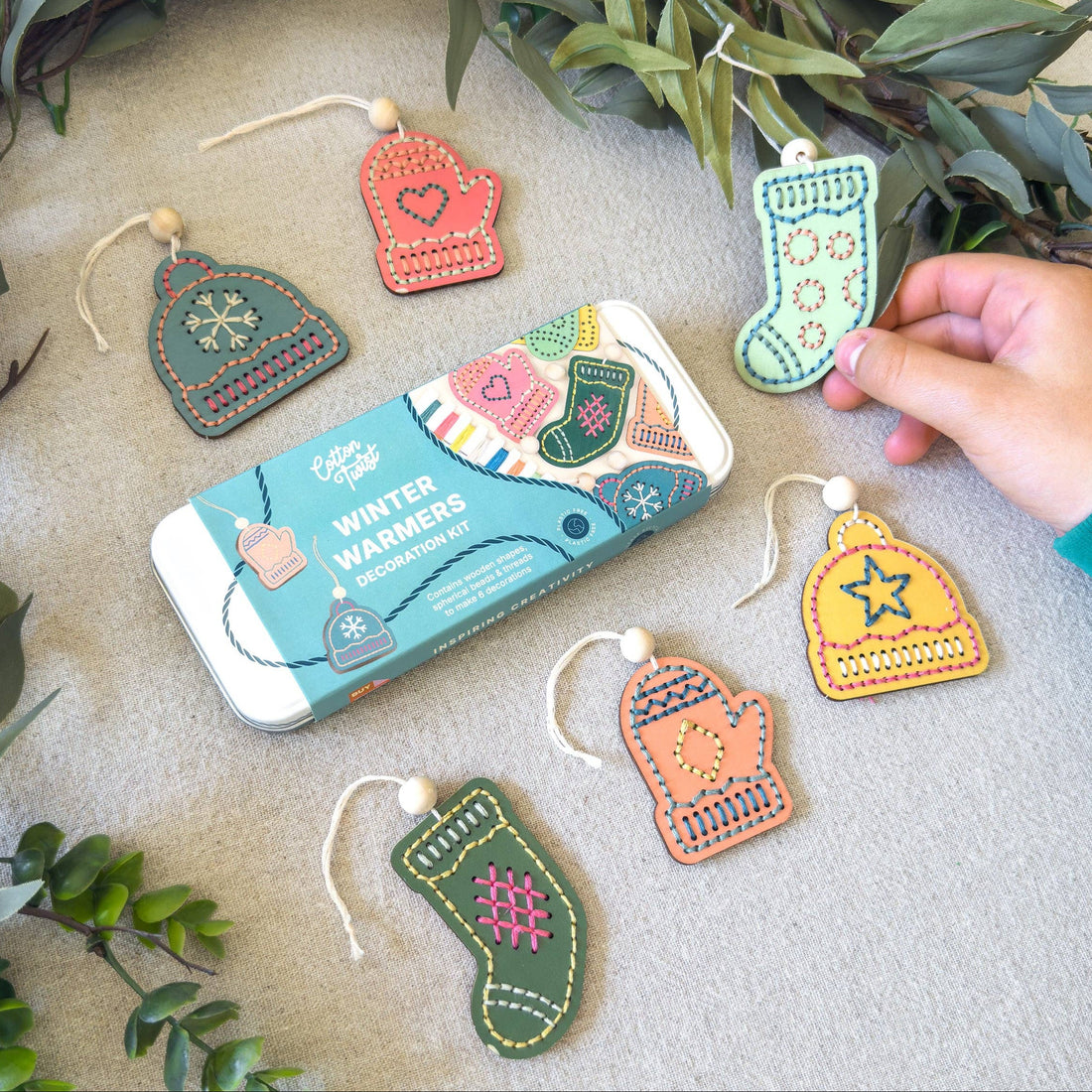 A tin of hand-embroidered wool felt Christmas ornaments is displayed on a neutral fabric surface with green foliage accents. A person's hand is holding one of the green stocking ornaments.
