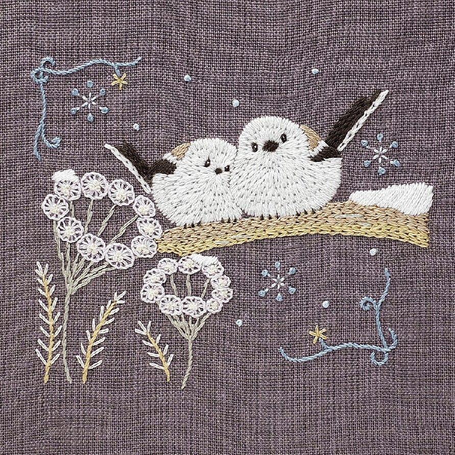 Embroidered design of two birds on a branch with flowers and snowflakes on a textured fabric background.