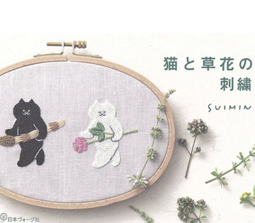Embroidery hoop with black cat and white cat design on fabric, surrounded by small plants.