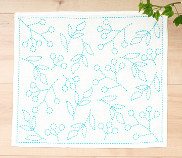 Embroidery pattern with blue thread on a white fabric square, placed on a wooden surface with green leaves.