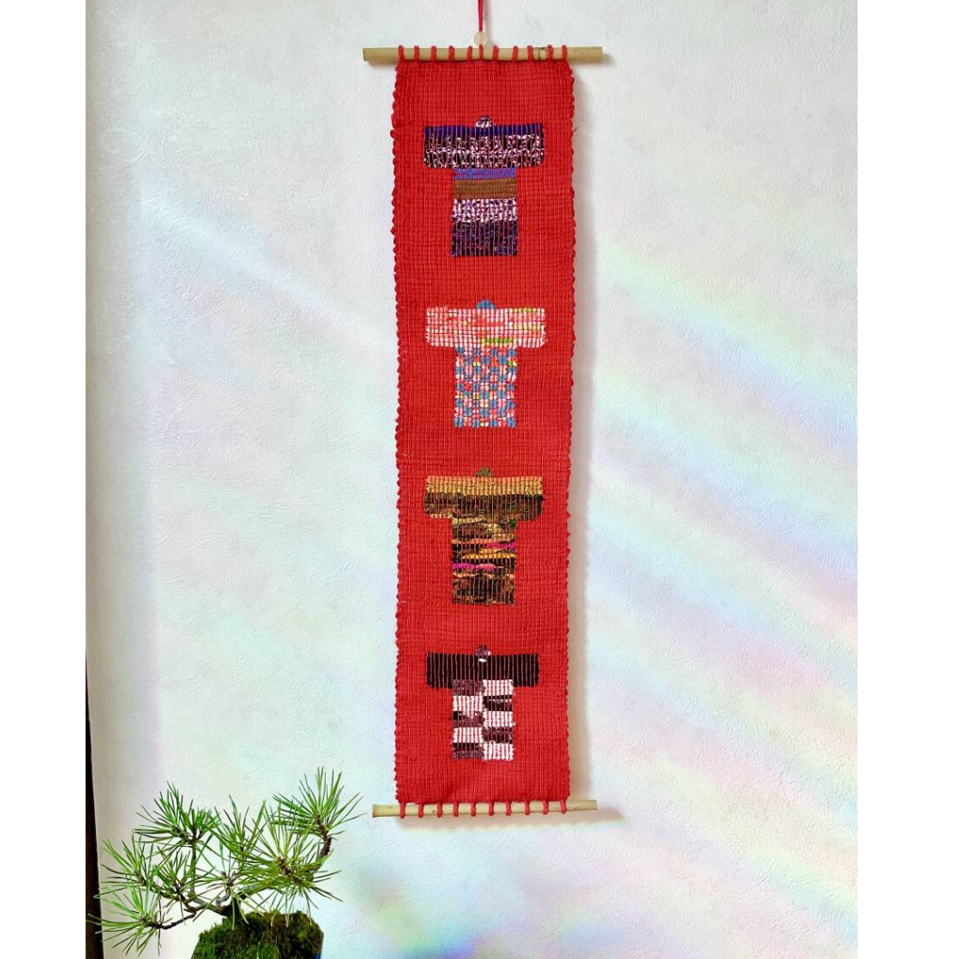 Textile art piece with colorful patterns on a red background, hanging on a white wall.