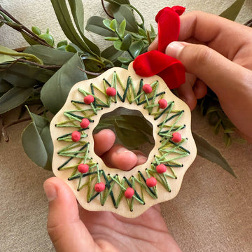 Handmade wooden ornament with greenery and red berries held by a person against a natural background.