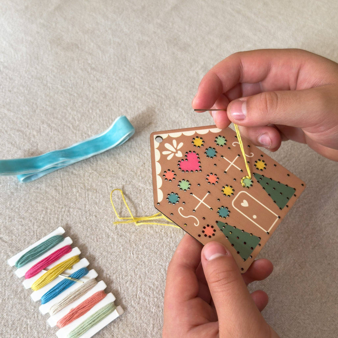 A person's hands are shown holding a wooden house-shaped embroidery kit over a beige fabric surface, with a blue ribbon and a small spool of multicolored thread nearby.