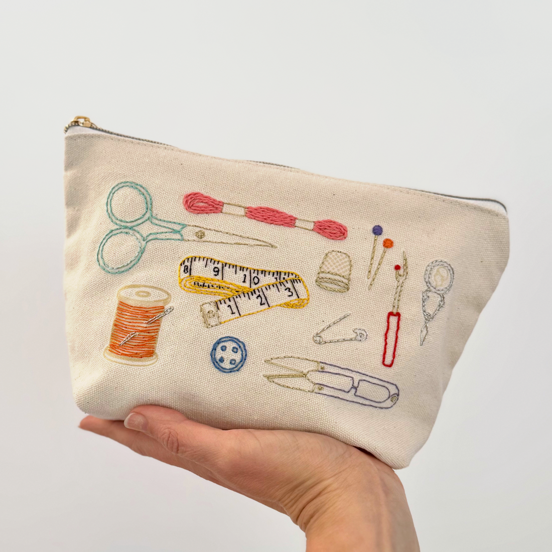 Hand holding a pouch with embroidered sewing tools on a light gray background
