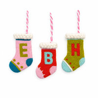 Three small felt Christmas stockings hanging on a white textured background, each with a pink-and-white striped loop. The stockings are pink, red, and olive green with white faux-fur trim and large letters E, B, and H in contrasting colors.
