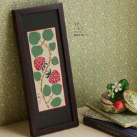 Framed cross-stitched pattern of berries and leaves on a textured green wall.