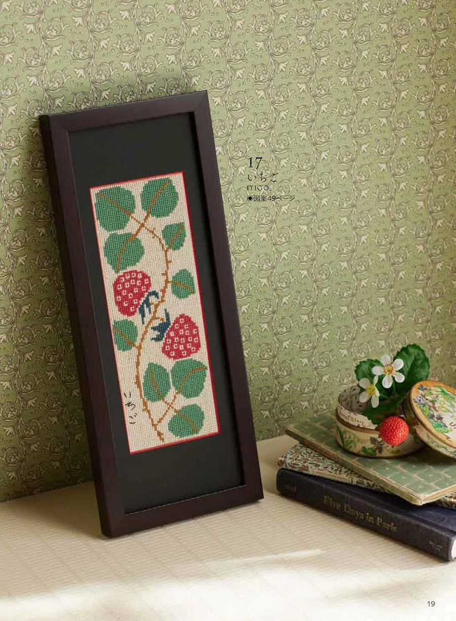 Framed cross-stitched pattern of berries and leaves on a textured green wall.