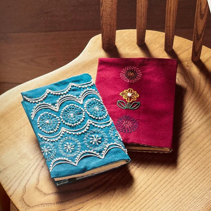 Embroidered fabric samples on a wooden chair with a magazine open to pages showing similar designs.